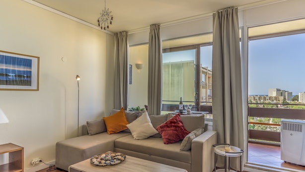 A modern living room with a grey sofa, a small side table, and a view of the city through large windows. - FLH Vilamoura Cozy Studio (Vilamoura)