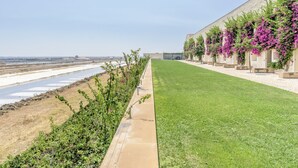 A hotel with a long, narrow building featuring a row of rooms with purple flowering vines. A well-maintained lawn runs alongside the building, and a body of water is visible in the background. - FLH Cozy Tavira with Pool (Tavira)