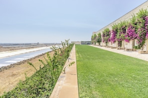 A hotel with a long, narrow building featuring a row of rooms with purple flowering vines. A well-maintained lawn runs alongside the building, and a body of water is visible in the background. - FLH Cozy Tavira with Pool (Tavira)