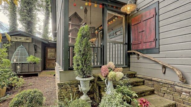 Une maison rustique en bois, dotée d’un escalier en pierre menant à une porte, et entourée de différentes plantes en pot ainsi que d’une nichoir.