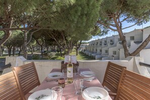A table set for dining with a bottle of wine, glasses, and plates, overlooking a row of white buildings and trees.
