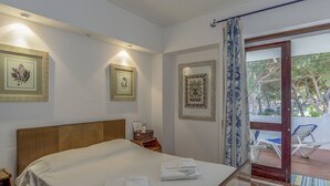A hotel room with a wooden bed, a nightstand, and a small table with a bottle and a basket. There are framed pictures on the wall and a view of the outdoor area with chairs. - FLH Albufeira Açoteias Apartment (Albufeira)