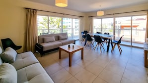 A modern living room with a sofa, a dining table with chairs, and large windows offering a view of the outside. - FLH Praia da Rocha Spacious Flat (Portimão)