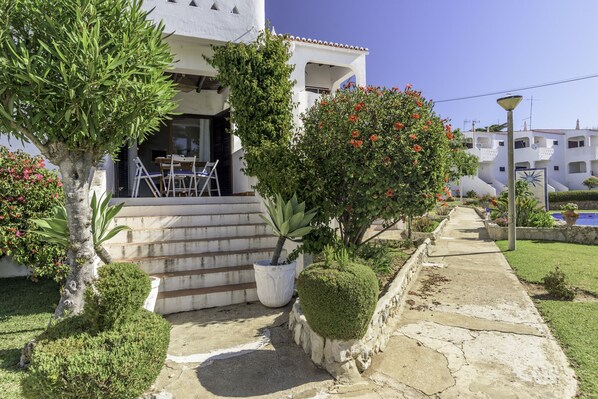 A white villa with a stone staircase leading to a covered patio, surrounded by well-maintained gardens and a clear blue sky.