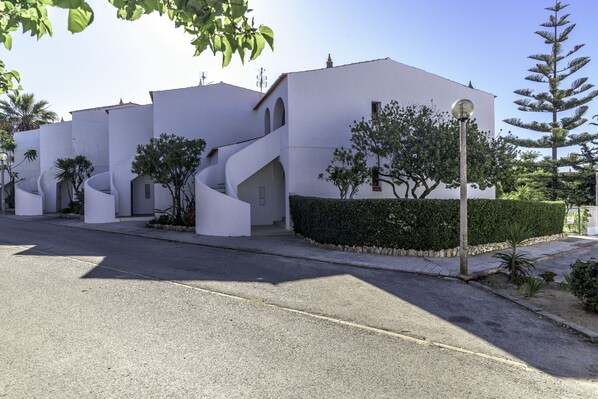 A modern building with a curved facade, a staircase leading to the entrance, and a well-maintained garden with various plants and a street lamp.