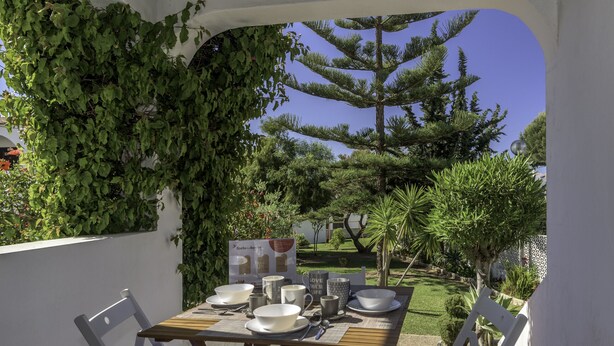 Uma área de refeições ao ar livre com uma mesa posta para quatro pessoas, rodeada por cadeiras brancas, sob uma pérgola coberta por trepadeiras verdes.