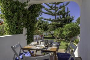 An outdoor dining area with a table set for four, surrounded by white chairs, under a pergola covered with green vines.