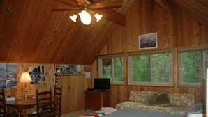 Uma cabana com interior de madeira, uma cama, uma mesa com abajur, uma televisão e uma janela com vista para árvores.