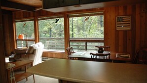 Interior de cabana com mesa de jantar, cadeiras, sofá e janelas amplas com vista para uma floresta.