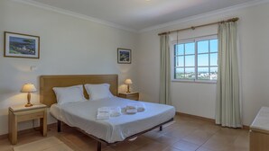 Un llit ben fet amb roba de llit i tovalloles blanques, un capçal de fusta, dues làmpades de nit, una finestra amb cortines i dos quadres al mur.