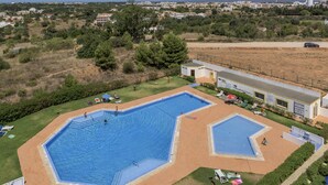 Una gran piscina exterior amb una piscina més petita a prop, envoltada d'una zona verda i un edifici amb teulada plana.
