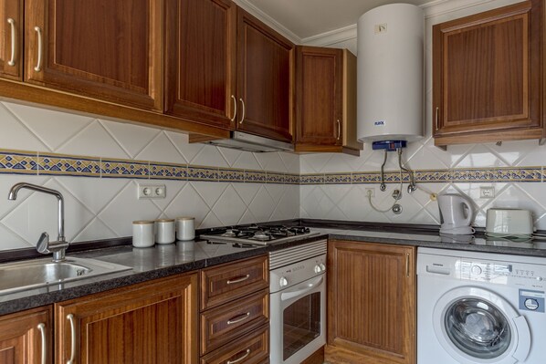 Een keuken met houten kastjes, een gasfornuis, een ingebouwde oven, een wasmachine en een boiler.