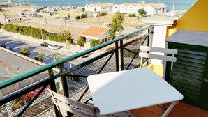 Un balcon avec une table et une chaise blanches, donnant sur un parking et offrant une vue sur la côte, avec des bâtiments et une étendue d’eau.