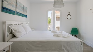 A white bedroom with a bed, a nightstand, a green chair, and a view of the outside. - FLH Altura Family House with Pool (Altura)