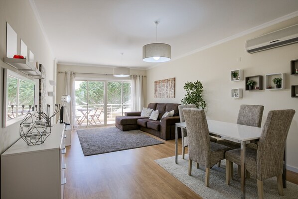 A modern living room with a sofa, dining table, and chairs. There is a large window with curtains, a plant, and wall-mounted shelves with decorative items.