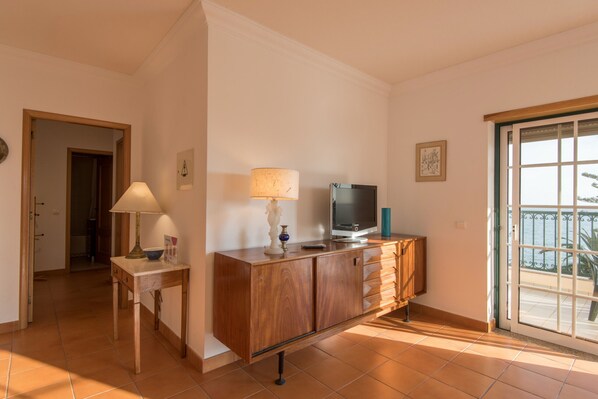 A room with a wooden cabinet, a television, a lamp, and a door leading to a balcony. - FLH Armação de Pêra Ocean View Flat (Armação de Pêra)