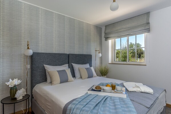 Un dormitorio ordenado con una cama grande, una mesita de noche con un florero y una ventana con vista a la vegetación.