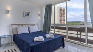 Ein Schlafzimmer mit Bett, Nachttisch, einem Fenster mit Vorhängen und Blick auf Gebäude.