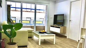 Een moderne woonkamer met een bank, een salontafel, een televisie en een groot raam met uitzicht op een balkon.