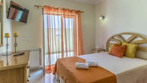 Una camera d'albergo con un letto, una TV fissata al muro, un comodino con una lampada e una finestra con tende leggere.