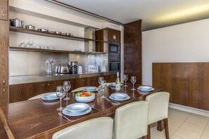 A modern dining area with a wooden table set for four, equipped with a built-in oven and microwave, and shelves above holding kitchenware.