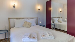 Un lit bien fait, avec du linge de lit blanc et deux serviettes, une table de chevet avec une lampe, et un grand miroir qui reflète la pièce.