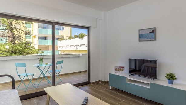 Un salon moderne avec une télévision, une petite table et un canapé. Il y a un balcon d’où l’on peut admirer les bâtiments et les arbres.