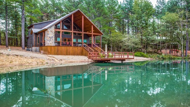 A cabin with a stone foundation, wooden deck, and a dock extending into a calm lake, surrounded by dense forest. - Enjoy Pier on a Pond, Hot Tub, Playset, Games, Outdoor Furniture, Fireplace, TV (Broken Bow)