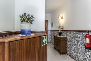 Ein Empfangsbereich mit Holztresen, einer Blumenvase, einem Feuerlöscher und Wänden mit gemusterten Fliesen.