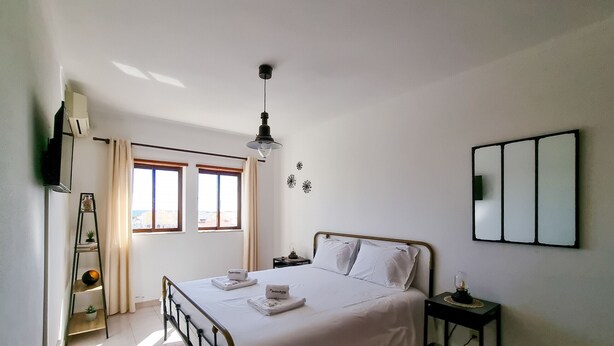 Un dormitorio con cama, mesitas de noche, televisión, ventana y espejo instalado en la pared.