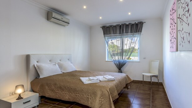 A bedroom with a bed, a nightstand, a chair, a window with curtains, and wall art. - FLH Albufeira Vila Mira Flat with Pool (Albufeira)