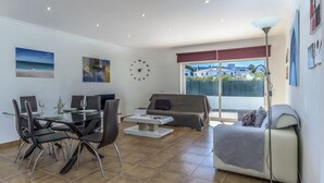 A modern living room with a glass dining table, black chairs, a beige sofa, and a large window with a view of buildings. - FLH Albufeira Vila Mira Flat with Pool (Albufeira)