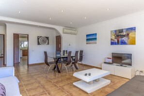 A modern living room with a glass dining table, comfortable seating, a flat-screen TV, and artwork on the walls.