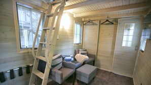 A cozy room with a sofa, two armchairs, a small table, and a ladder. There are two windows with blinds, a door, and a hanging light fixture.