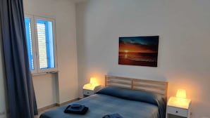 A bedroom with a bed, two bedside tables with lamps, a window with blue shutters, and a sunset painting on the wall. - Ville Ninfea - 2 independent twin pool villas, 150 mt by the sea Fontane Bianche (Fontane Bianche)