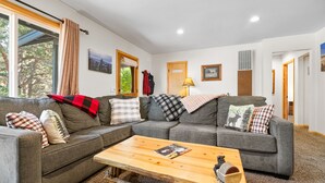 Ein Wohnzimmer mit einem grauen Ecksofa, einem Holztisch und Blick auf Bäume durch ein Fenster.