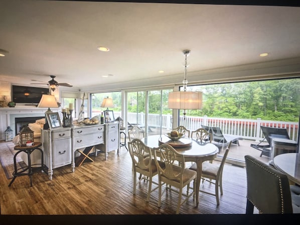 A spacious room with a wooden floor, a dining area with a table and chairs, a fireplace, and a large window overlooking a deck.
