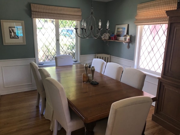 A dining room with a wooden table, white chairs, a chandelier, and a window with a view of greenery.