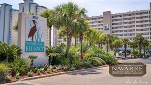 Exterior del hotel Navarre Beach Regency con un letrero que muestra un pelícano y palmeras, rodeado de exuberante jardinería.