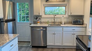A modern kitchen with white cabinets, stainless steel appliances, and a window with a view of greenery. - Late August and fall dates available! (Dennis)
