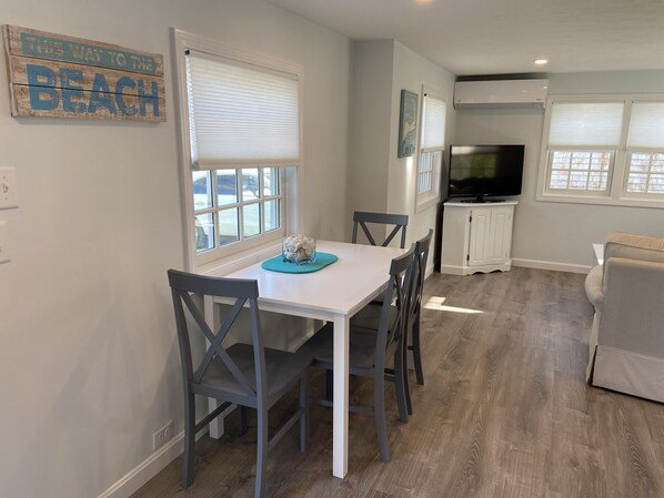 A dining area with a white table and gray chairs, a kitchen with a TV and white cabinets, and a living area with a sofa.