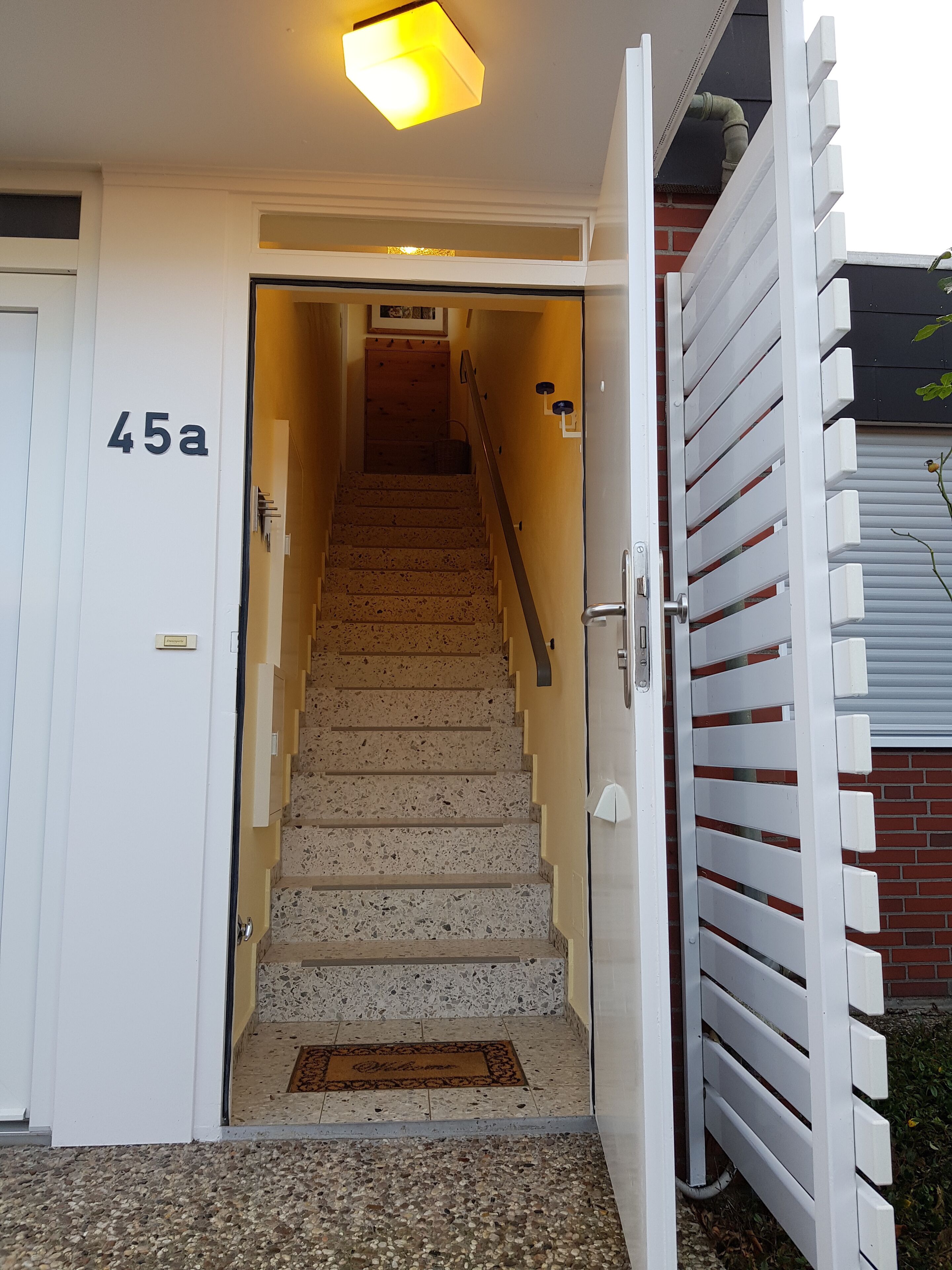 Un escalier menant à une porte marquée du numéro « 45a », devant laquelle se trouve un paillasson sur lequel est écrit « Bienvenue ».