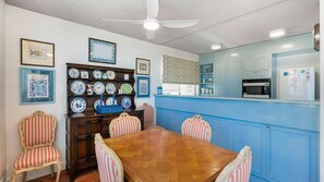 A dining area with a wooden table and striped chairs, a blue kitchen with a refrigerator, and a ceiling fan.