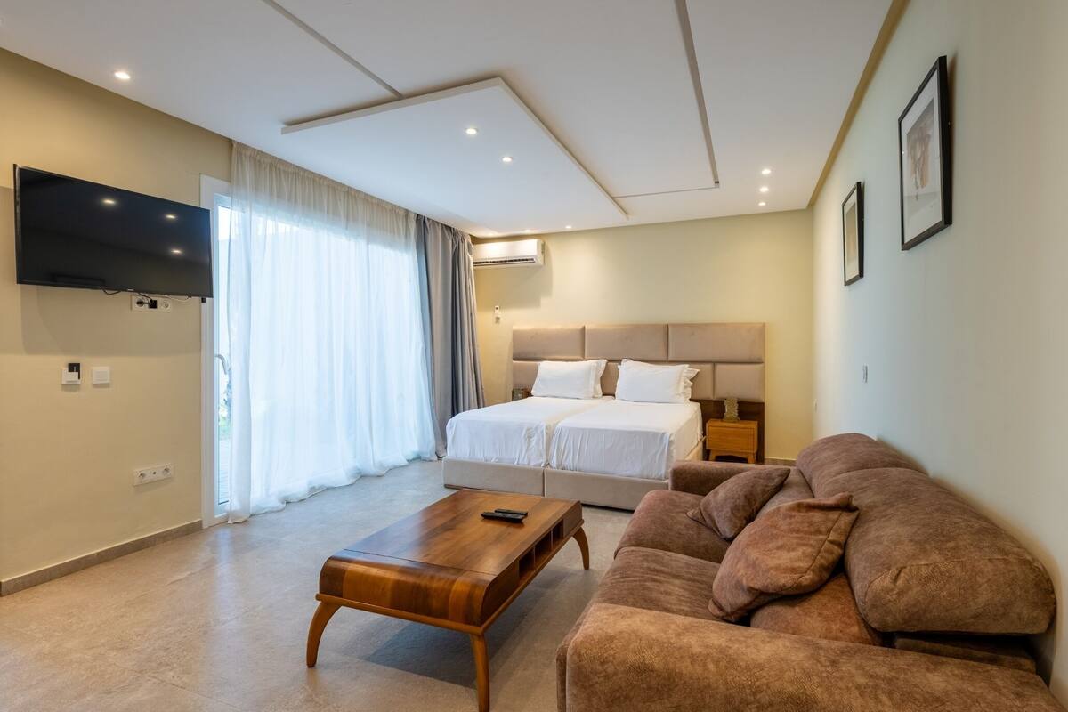 Una habitación de hotel con una cama grande, un sofá, una mesa de centro de madera, un televisor montado en la pared y una ventana con cortinas.