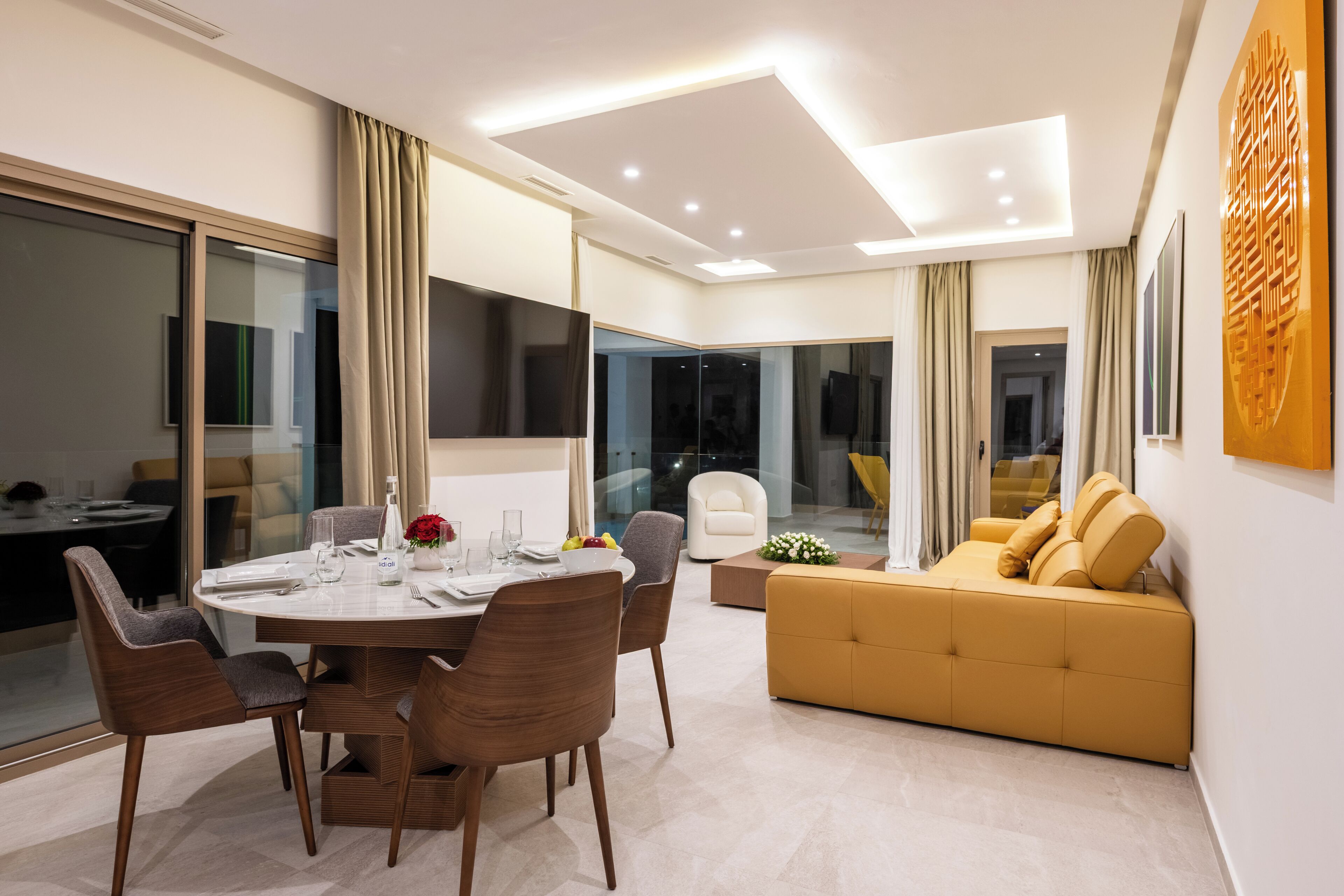A modern living room with a round dining table, brown chairs, a yellow sofa, and a large flat-screen TV.
