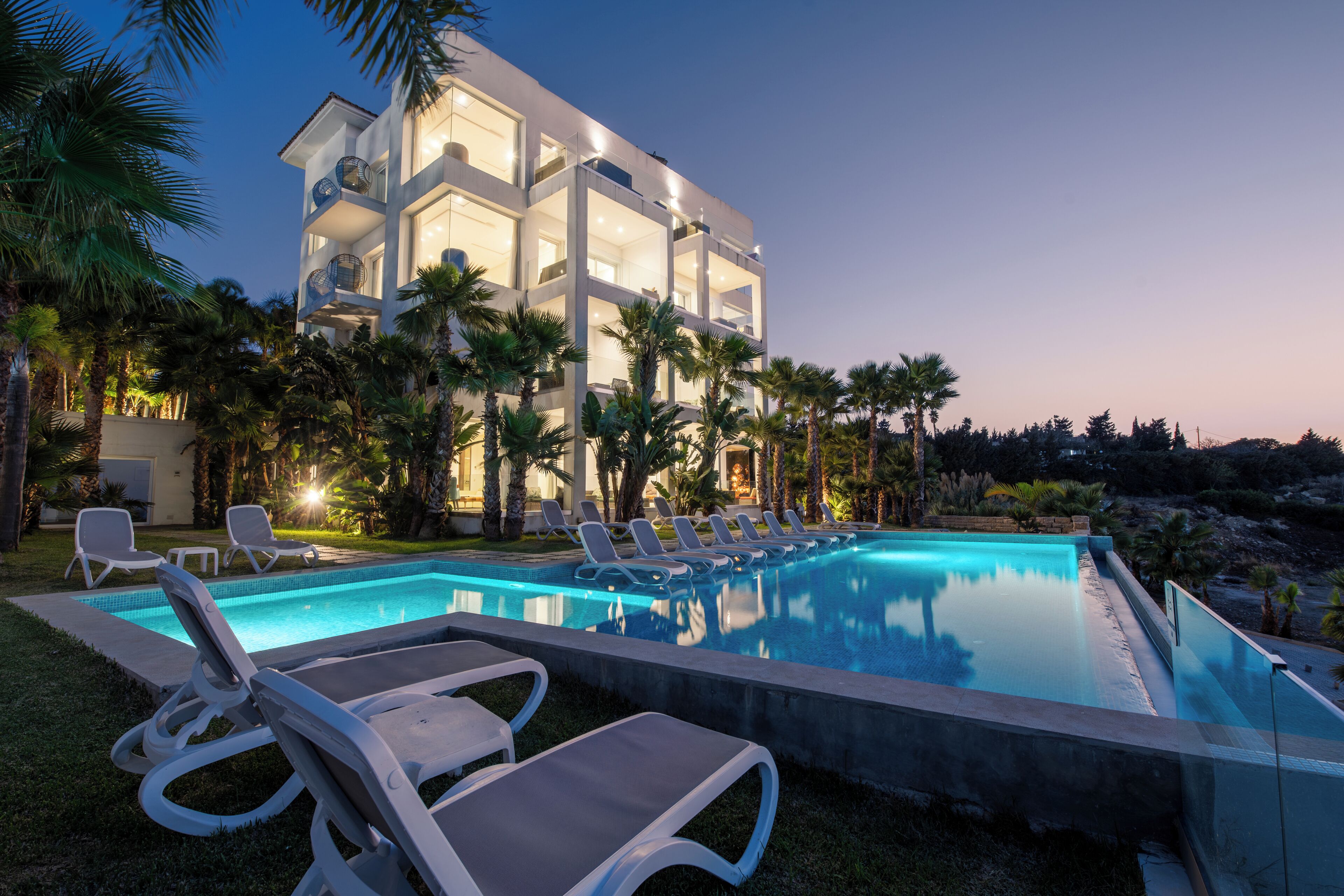 A modern hotel with a swimming pool, sun loungers, and palm trees.