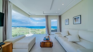 A spacious living room with a large window offering a view of the sea and greenery. There is a white sofa, a wooden coffee table, and a flat-screen TV on a wooden stand. - Le tanger (Al Bahraoyine)