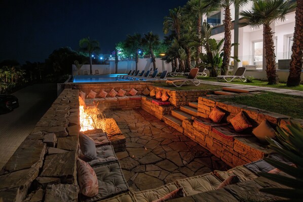 Un foyer en pierre avec des coussins, une piscine et des chaises longues, le tout installé dans un espace extérieur bien éclairé, pour une ambiance agréable le soir.