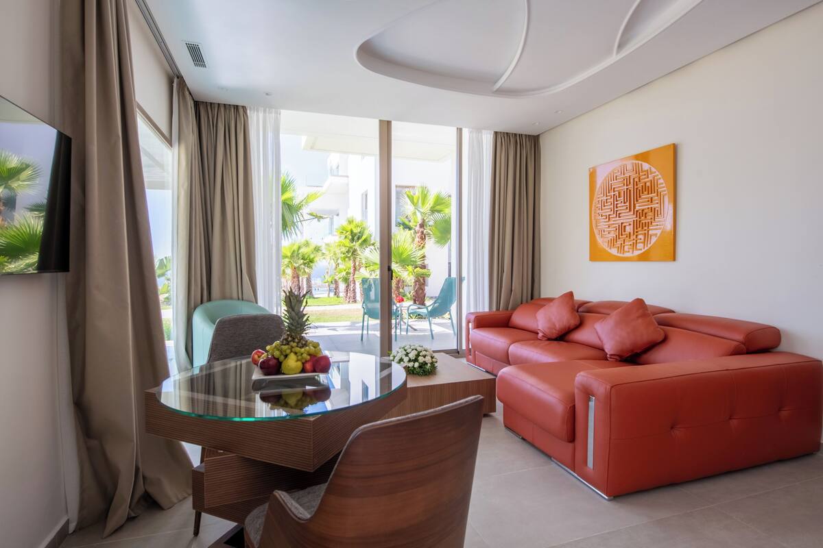 Una sala moderna con una mesa de comedor de cristal, un sofá rojo y vistas a un jardín tropical a través de grandes ventanales.