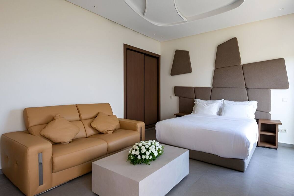 Una habitación de hotel moderna con un sofá de cuero marrón, una cama grande con cabecera y una mesita de noche pequeña.